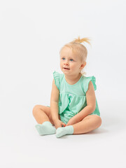 A sweet bright girl of 2 years old is sitting and attentively enthusiastically looking up at a white background in socks and a green dress. Expressive look.