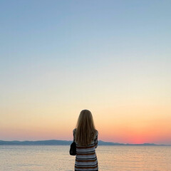 rear view of woman standing alone looking out to sea against sunset sky with space for text - obrazy, fototapety, plakaty