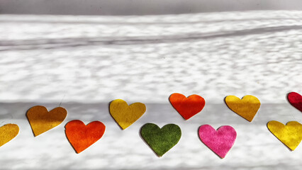 Multicolored hearts made of rough velvet paper on a white surface with stripes of shadow and light from the sun. Abstract background, texture, copy space