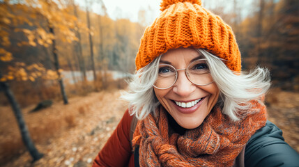 Happy older woman smiles takes selfie in autumn park. Positive cheerful woman in orange hat enjoying walk outdoors - obrazy, fototapety, plakaty