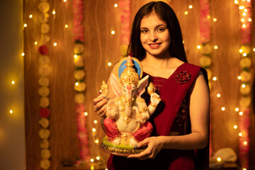 Indian woman holding Ganesh idol or Ganesh Murti with festive lights and flower decoration in the...