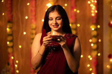 Portrait of a beautiful young smiling Indian woman in traditional dress holding oil lamp light or diya with festive lights decoration in the background celebrating diwali.