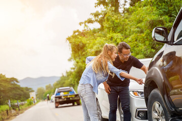 A female driver and a male driver sufferer point out the damage on car after accident on roadside with sun flare background.