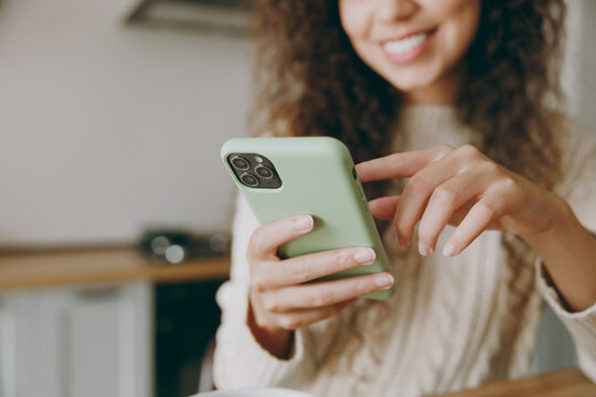 Cropped young smiling housewife woman wearing casual clothes sweater hold use mobile cell phone drink coffee look for recipe sit at table in light kitchen at home alone Lifestyle cooking food concept