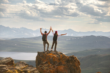 Young couple of hikers with backpacks are standing at mountain top with open arms in winner pose