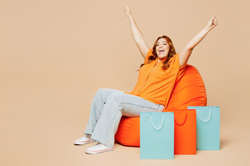 Full body young winner fun overweight woman wear casual clothes hold shopping paper package bags sit in bag chair isolated on plain beige background studio portrait. Black Friday sale buy day concept.