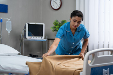 Indian nurse or caregiver preparing hospital ward bed by arranging pillow and blanket for patient -...