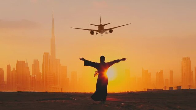 Back view of Arab woman rises her hands on the sun set. Passengers Airplane preparing for landing at Dubai International airport. Dubai city on the background.