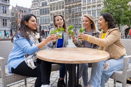 A lively image featuring a multi-ethnic group of female friends savoring refreshing Mojito cocktails on a cafe terrace located in a bustling European city center. The scene encapsulates camaraderie