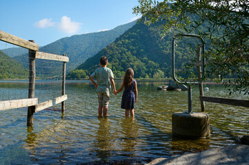 Brother and sister holding hands, standing close to each other barefoot in Segrino lake in Lombardy in Italy. Rear view - Powered by Adobe