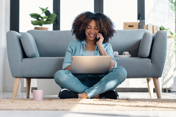 Obraz premium Pretty young woman talking with smartphone while working with laptop sitting on the floor at home.