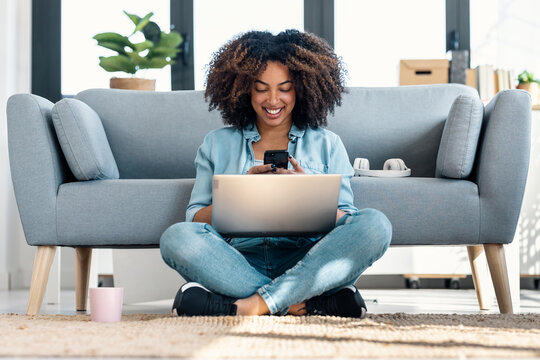 Pretty young woman using her mobile phone while working with laptop sitting on the floor at home.