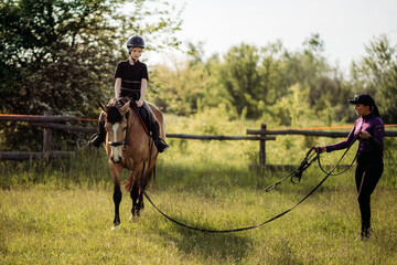 A teenage boy in a helmet learns horse riding in the summer, the instructor teaches the child equestrianism.