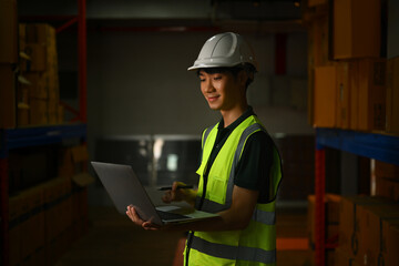 Warehouse manager wearing hardhat and reflective jacket using laptop for checking stock and order details in warehouse