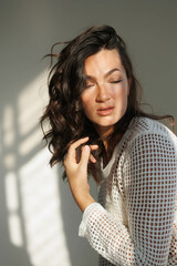 Beautiful brunette woman in white net top and blue jeans, sitting. Girl smile, happy. Portrait of young pretty woman. Complicated sunlight with shadows. White background. Light from window, portrait.