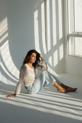 Beautiful brunette woman in white net top and blue jeans, sitting. Girl smile, happy. Portrait of young pretty woman. Complicated sunlight with shadows. White background. Light from window, portrait.