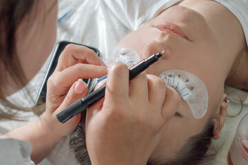 Lashmaker marking places for gluing artificial eyelashes on patches on lower eyelids of young woman with closed eyes.