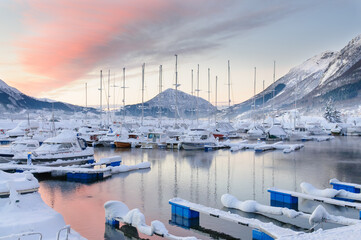 Winter sunrise over snow-covered marina with moored boats and mountainous backdrop