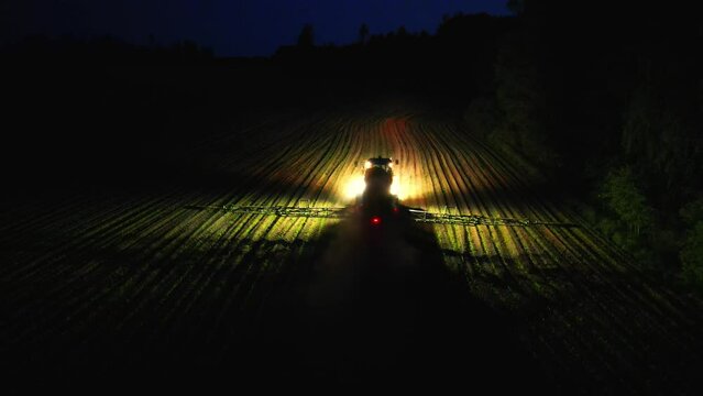 Tracktor in field after sunset. Aerial view of harvesters spraying pesticide. Aerial view of combine harvester working at night. Illuminated field at night during the end of harvesting season