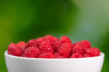 Beautiful ripe juicy raspberries in a white bowl on a green blurry background, macro photography,...