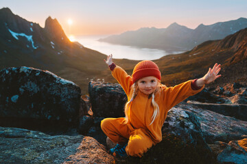 Child girl hiking in mountains travel vacations in Norway adventure outdoor active healthy lifestyle 4 years old kid happy raised hands enjoying midnight sun landscape - obrazy, fototapety, plakaty