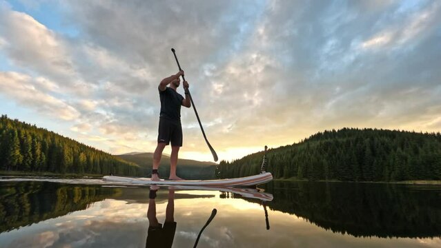 Water reflection of man silhouette standing on stand up paddle board doing sup water sport activity on a mountain lake at sunset. Healthy lifestyle outdoor activity 