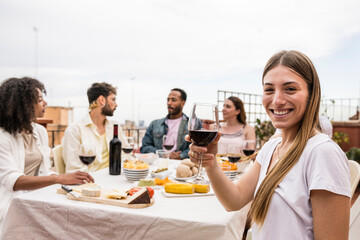 Young cheerful lady looking at camera with a glass of wine in a rooftop dinner party. Joyful beautiful woman toasting and staring at camera at outdoors terrace.