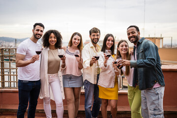 Diverse group of friends having drinks and toasting in a rooftop party.Multiracial young happy group of millennials drinking wine in a terrace looking at camera.