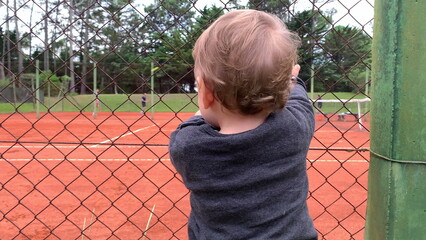 Cute baby holding into tennis court fence looking at siblings play game