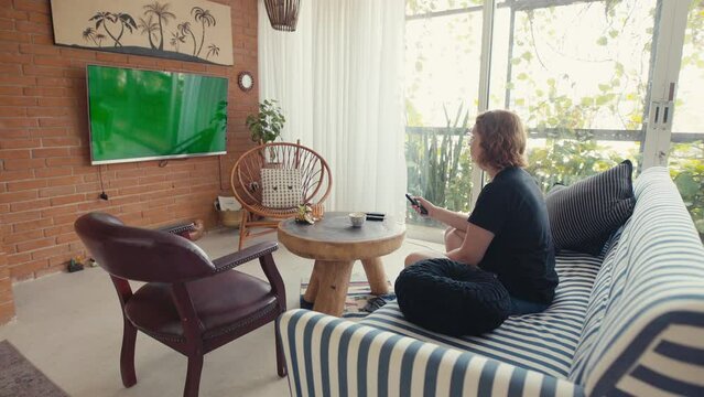 Casual woman turning on the TV in chroma key screen and settling on the sofa at home.