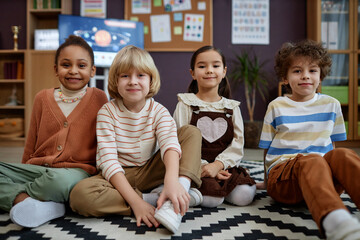 Front view of multiethnic group of little kids sitting on floor in preschool class and looking at camera