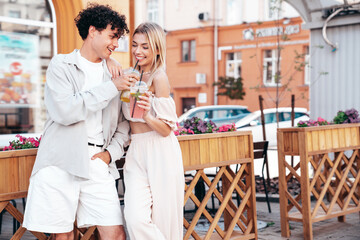 Young smiling beautiful woman and her handsome boyfriend in casual summer clothes. Happy cheerful family. Female having fun. Couple posing in street. Holding and drinking cocktail drink in plastic cup