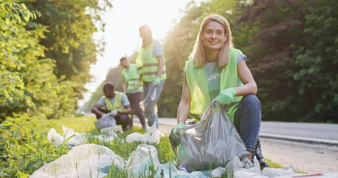 Portrait of young and attractive Caucasian woman cleaning up area near road with other volunteers. Enthusiastic and diverse group of people working together picking up trash helping nature. Pollution.