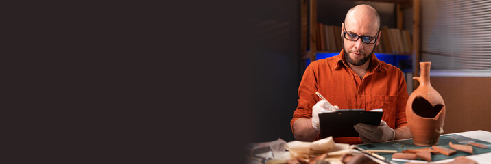 Bearded archaeologist in glasses working in the office late at night making notes on a clipboard...