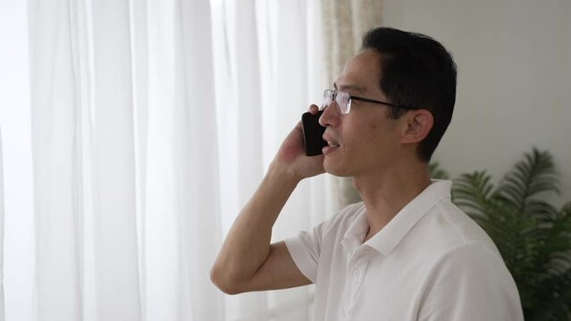 portrait of cheerful asian Chinese mature mid age man talking to friend on the mobile phone near window at home during the day. happy andropause and communication concept