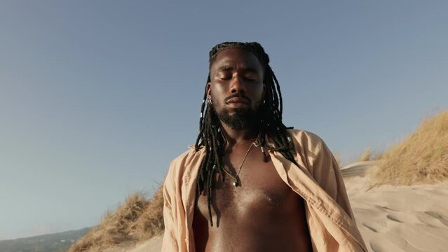 An African black male with stylish dreads and a loose open shirt, exudes confidence while turning to the camera against a sandy dunes background