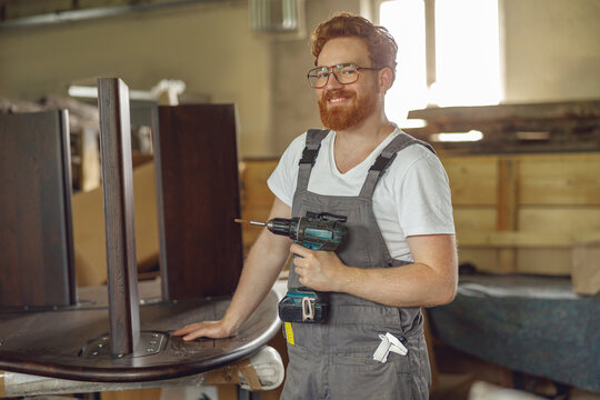 Smiling carpenter fixing table with electric drill standing in carpentry workshop and looks camera