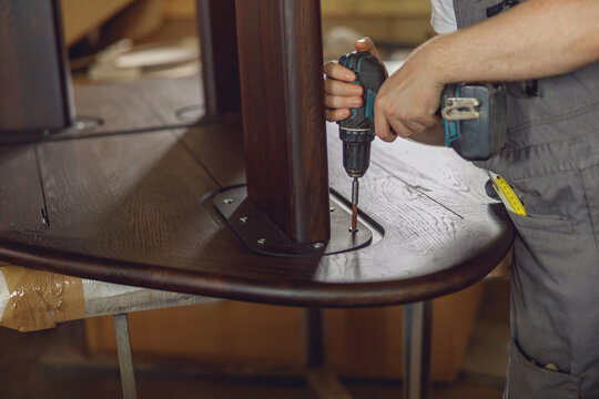 Close up of carpenter fixing table with electric drill standing in carpentry workshop and looks away