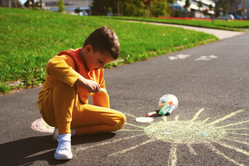 A little seven-year-old boy in an orange tracksuit and white sneakers draws the sun with colored chalk on the asphalt