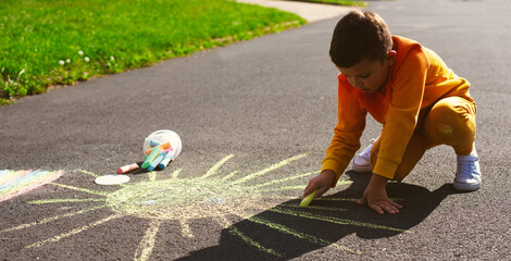 A little seven-year-old boy in an orange tracksuit and white sneakers draws the sun with colored chalk on the asphalt