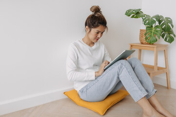 Fototapeta premium Happy asian Thai woman bun hair, sitting on floor, using tablet, working at apartment in winter day.