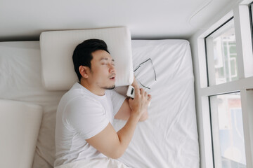 Asian man asleep, sleeping on white bed with phone in his hand and eyeglasses, snoring at apartment.