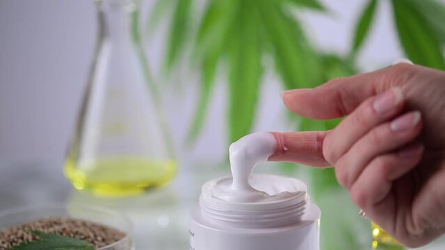 Female hand checking cannabis hemp cream. Container with hemp skin care cream, CBD oil, THC tincture and cannabis leaves. Cannabis medical herbal extract. Organic skin care CBD oil cream.