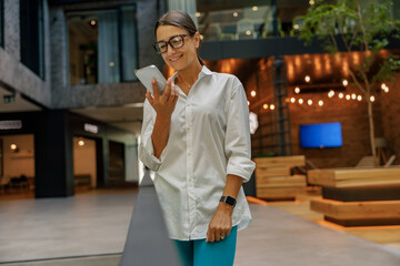 Female entrepreneur in eyeglasses looking on phone while standing in modern coworking space