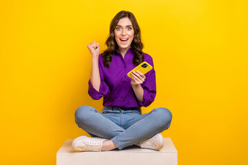 Full body photo of delighted crazy lady sit podium cube hold smart phone raise fists isolated on yellow color background