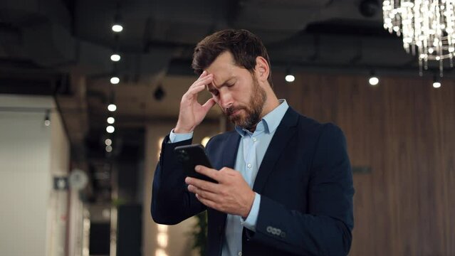 Astonished caucasian businessman receiving notification on smartphone about company's downgrading shares. Man dressed in formal wear feeling distraught and desperately touching head with hand.