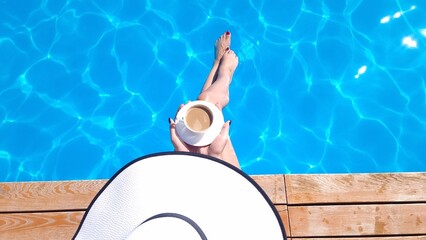 A girl in a hat sits near the pool with a cup of coffee