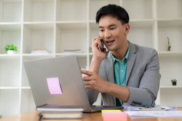 Asian businessman using smartphone and working at office desk using laptop and talking on mobile phone, financial business concept