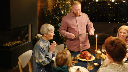 High angle of mature Caucasian man bringing roasted turkey to festive table for Christmas dinner and starting to cut it