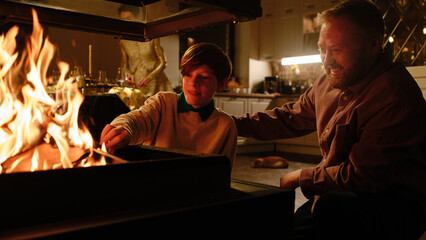 Mature Caucasian man and his son placing firewood into modern fireplace in living room at home